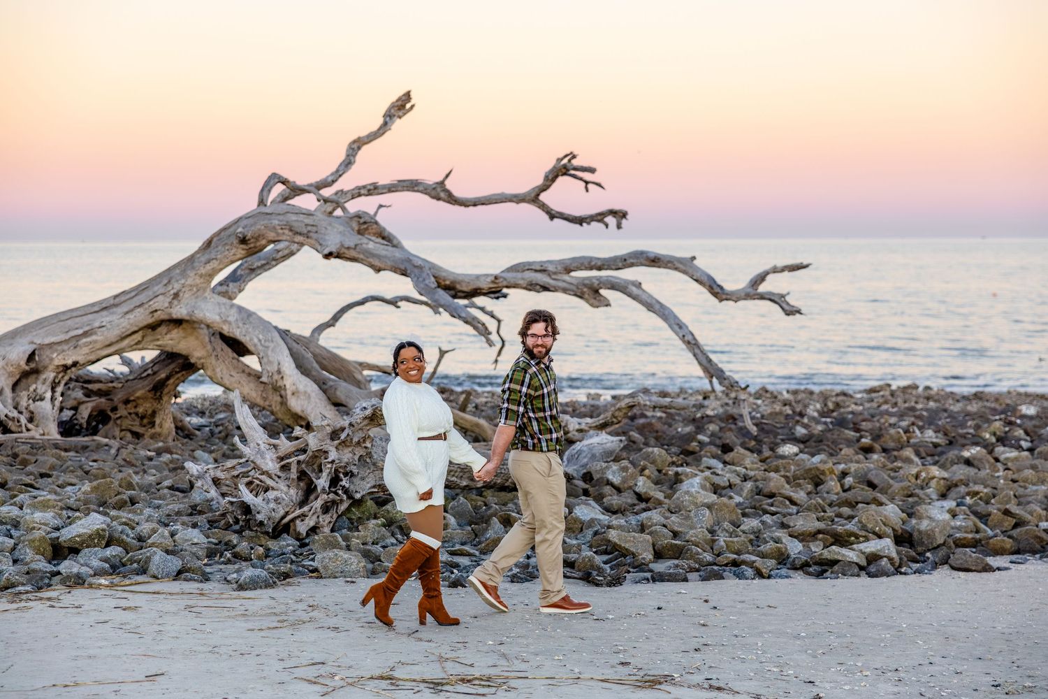 Engaged man and woman walking hand-in-hand on Driftwood Beach at sunset.
