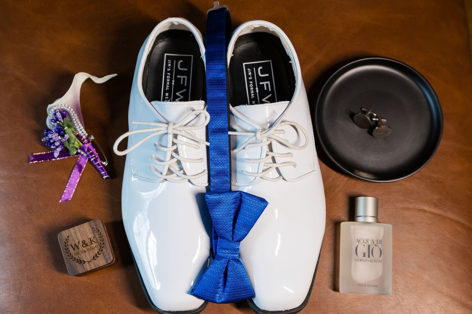 White patent leather dress shoes with blue bow tie, boutonniere, cologne, and ring box for a wedding groom.