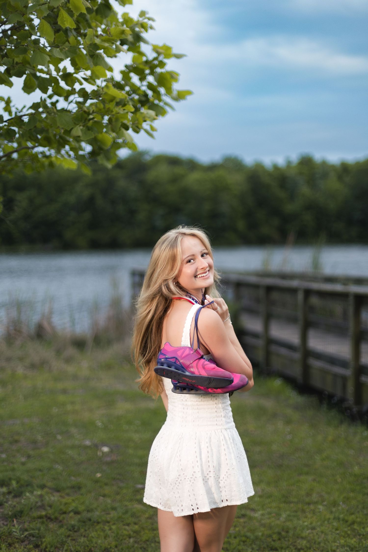 Smiling blonde teen in white dress holding colorful scarf near a lake and wooden bridge.