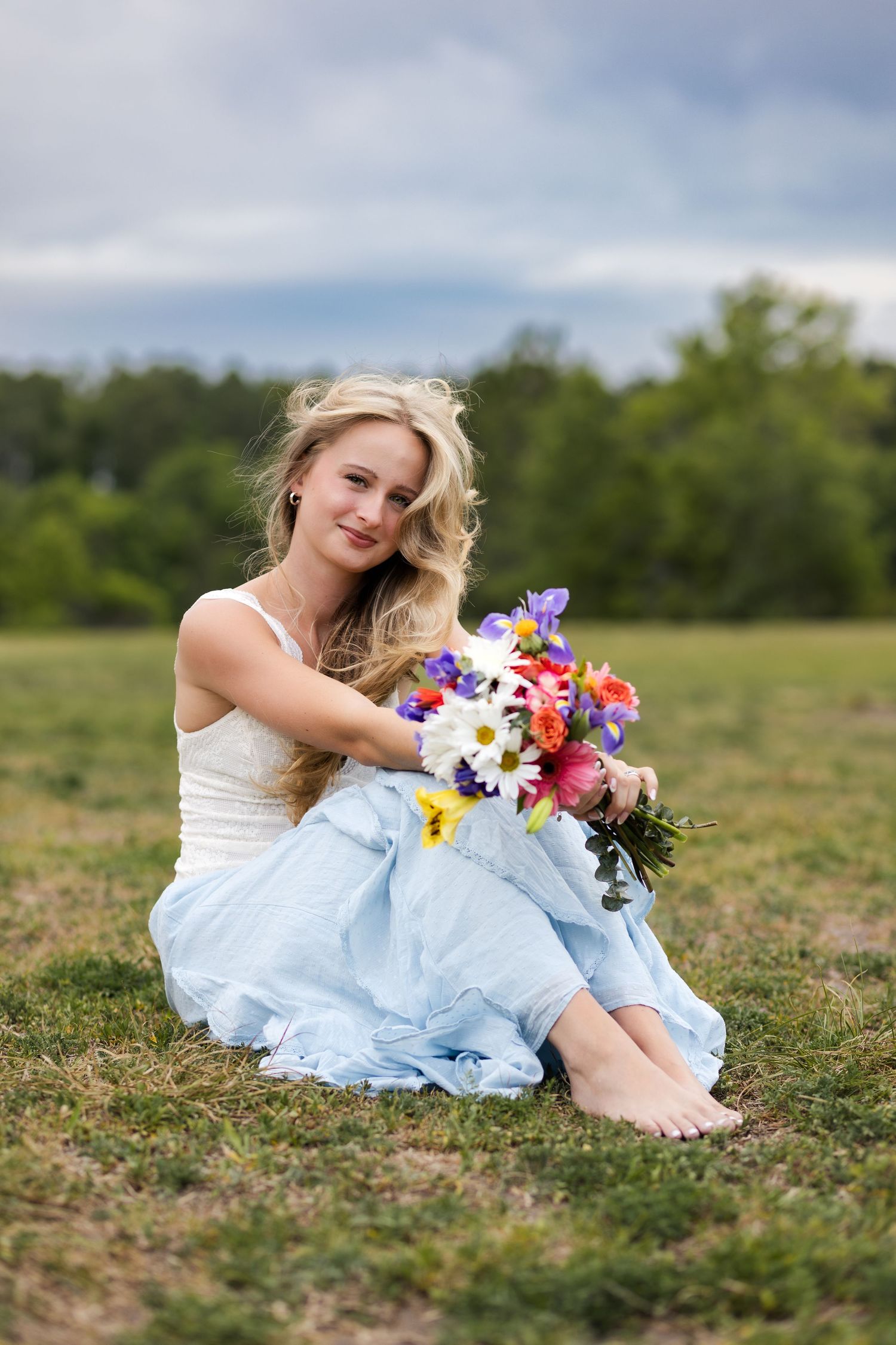 Smiling blonde woman sits barefoot on grass holding vibrant bouquet of mixed flowers in blue skirt.