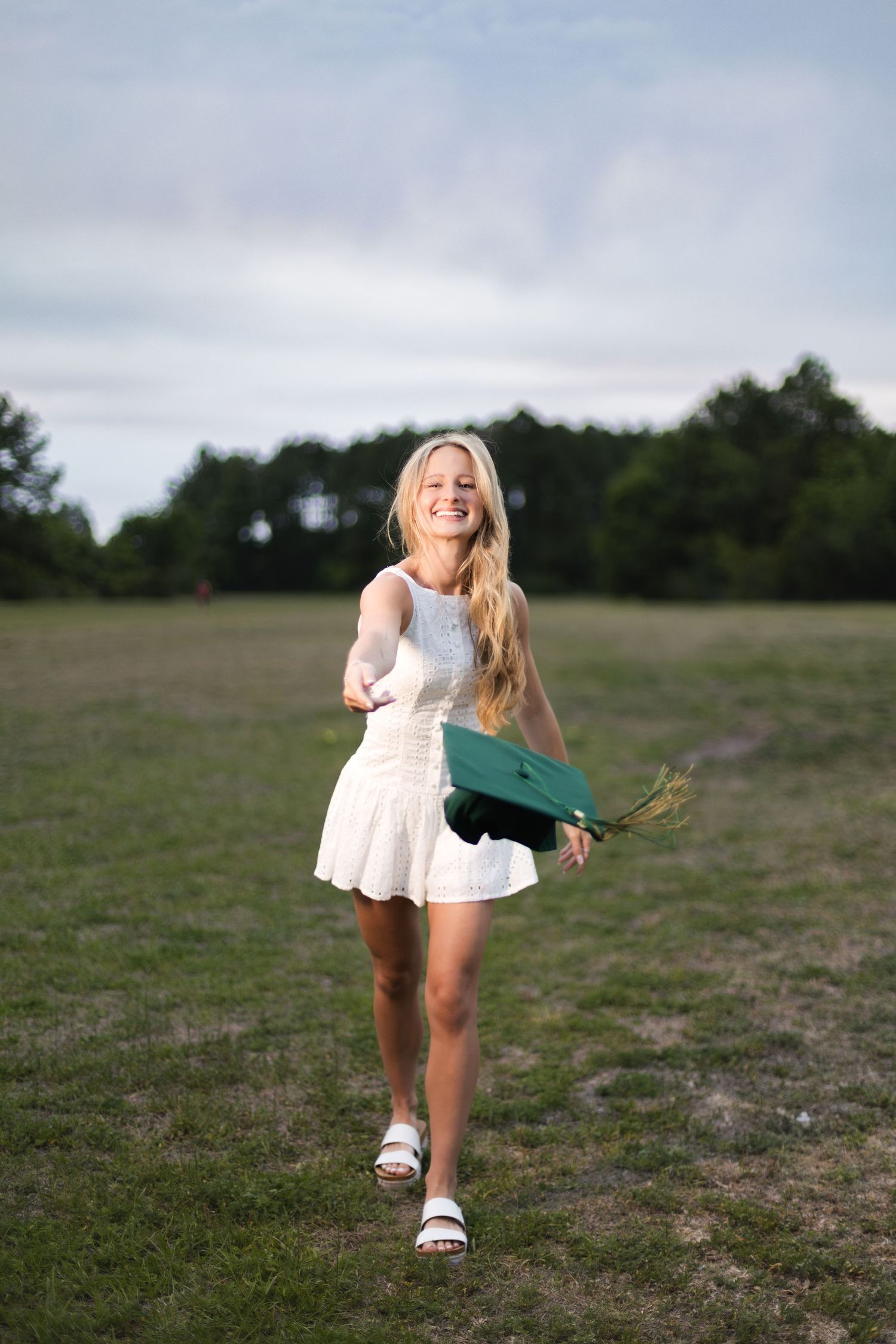 Happy blonde graduate in white dress holding green graduation cap while walking in an open field.