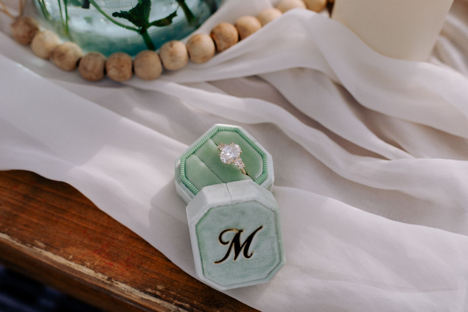 Elegant engagement ring displayed in a mint green velvet monogrammed ring box on a white fabric with wooden beads.