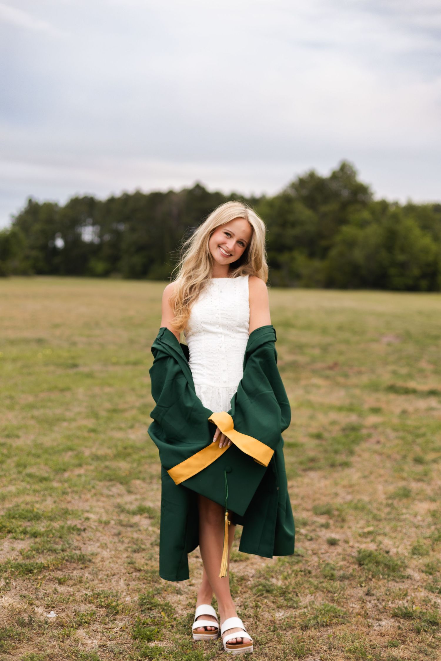 Blonde graduate smiling in white dress with green and gold graduation gown draped over shoulders outdoors.