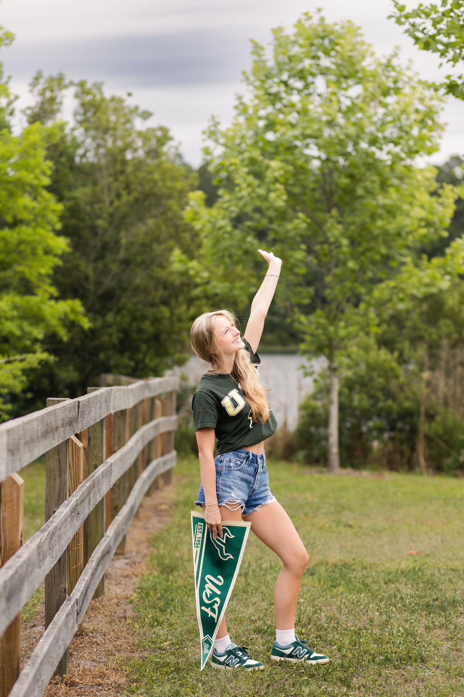 Blonde teenage girl holding a USF pennant flag beside a wooden fence in a green outdoor setting.