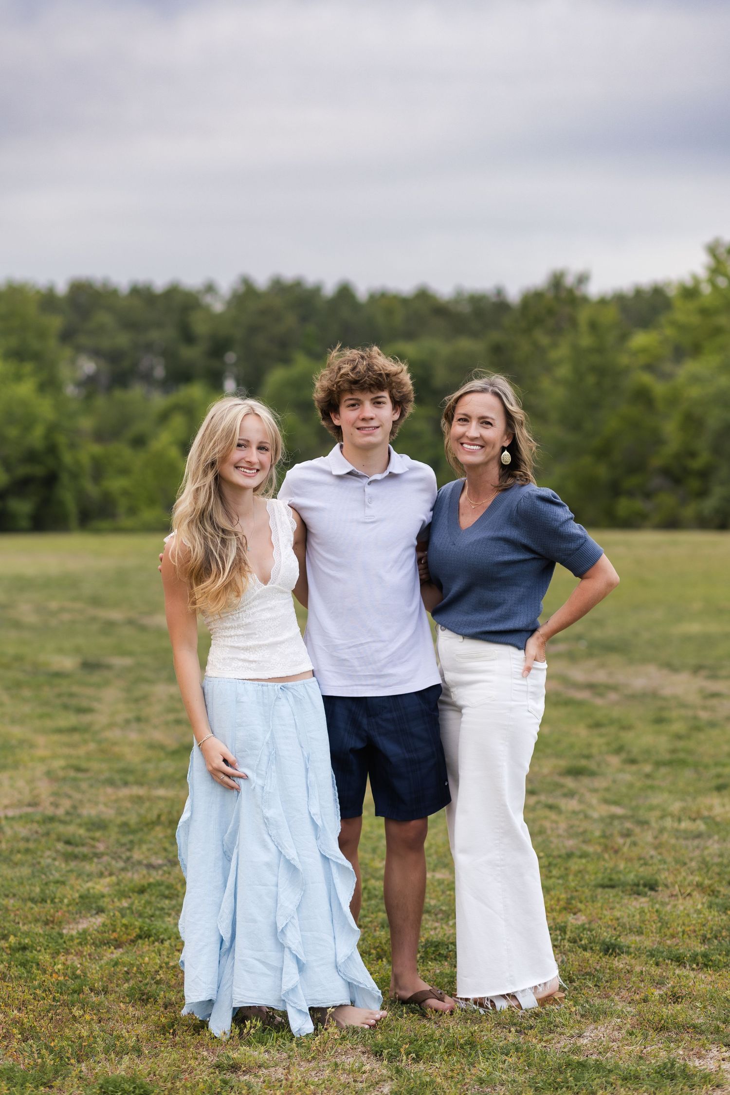 Family portrait of teenage girl, teenage boy, and mother smiling together in outdoor green field.