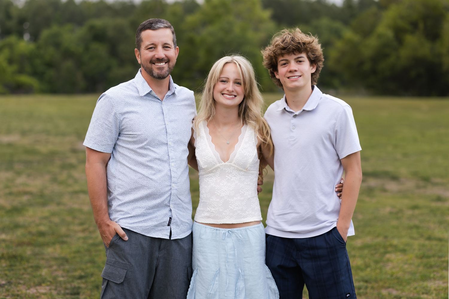 A smiling family of three poses outdoors on a green field, featuring a man, a blonde woman, and a teenage boy.