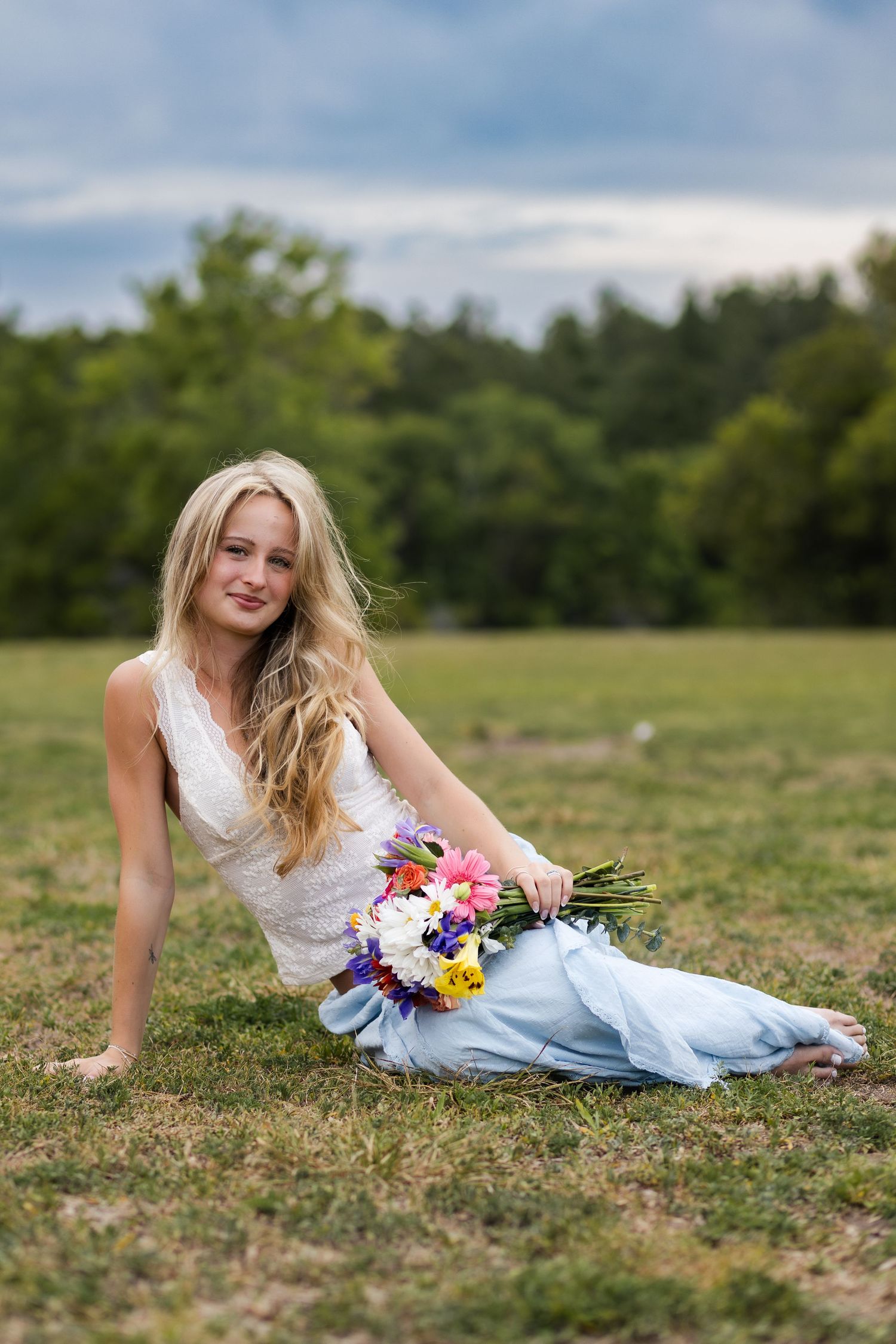 Blonde woman in white top and blue skirt sits on grass holding colorful wildflower bouquet outdoors.