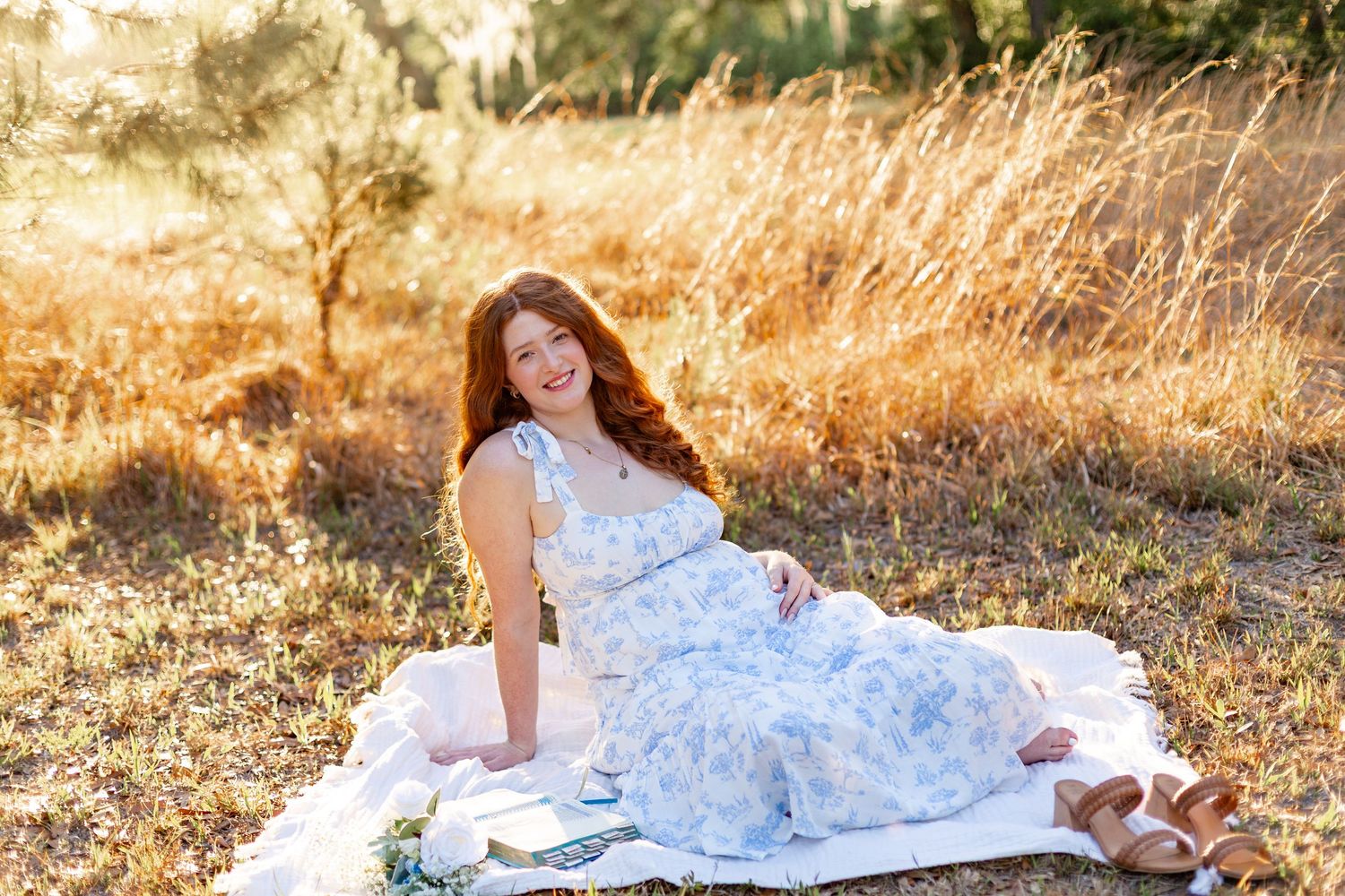 A smiling redhead woman in a blue floral dress sits on a white blanket in a sunlit golden field.