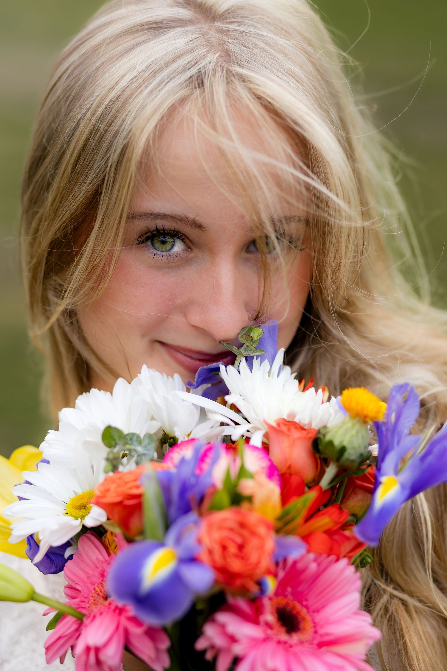 Close-up portrait of smiling blonde woman with green eyes holding colorful flower bouquet near face.