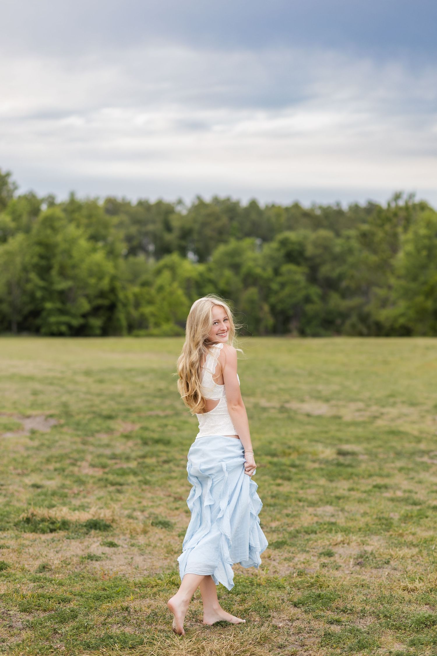 Blonde girl in white top and light blue ruffled skirt walking barefoot in a green field.