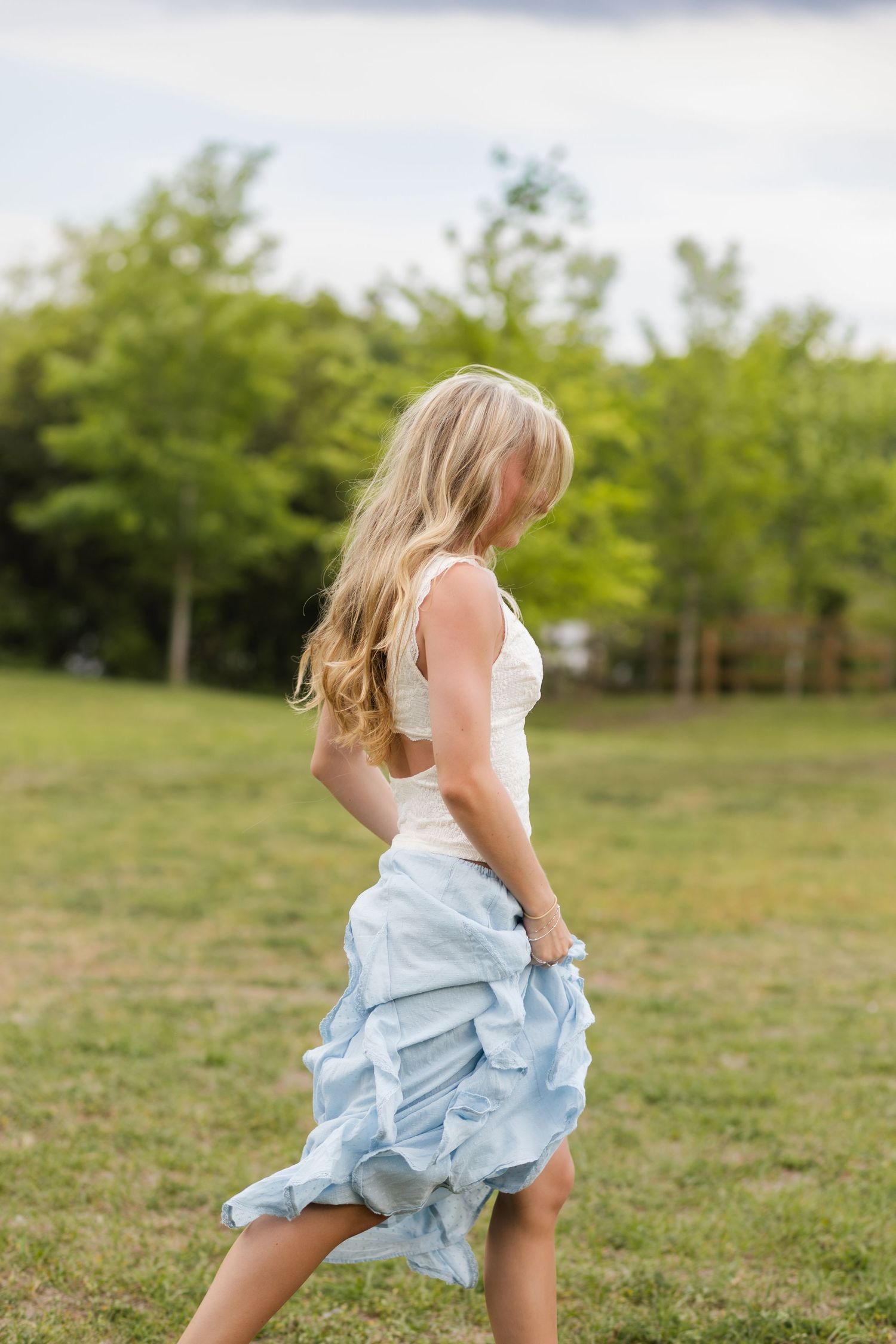 Blonde woman in white top and light blue ruffled skirt walking outdoors in a green grassy field.