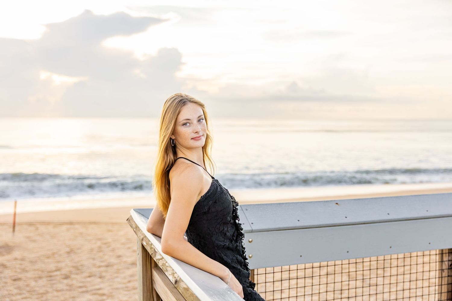 Senior girl smiling for photo, leaning on boardwalk rail during sunrise on Ponte Vedra Beach with ocean waves behind her.