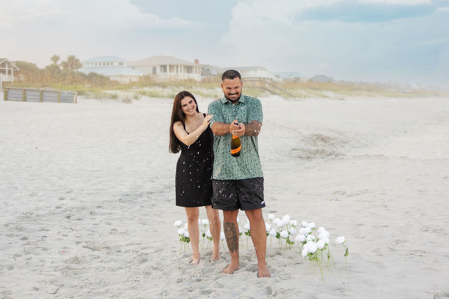 Couple celebrating beach proposal with champagne, flowers on white sand, wearing black dress and teal shorts.
