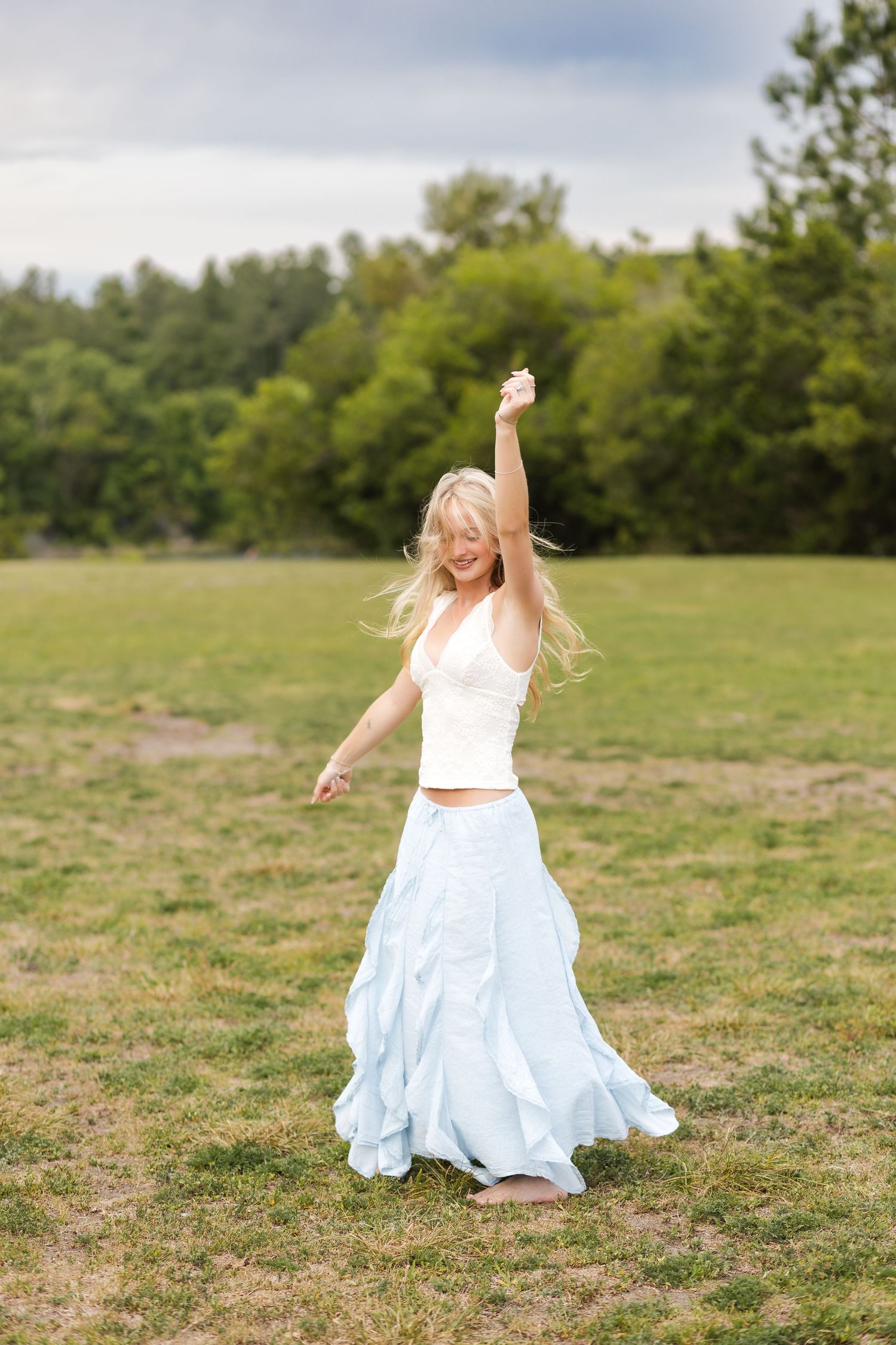 Joyful blonde woman dancing in a field wearing a white top and light blue maxi skirt.