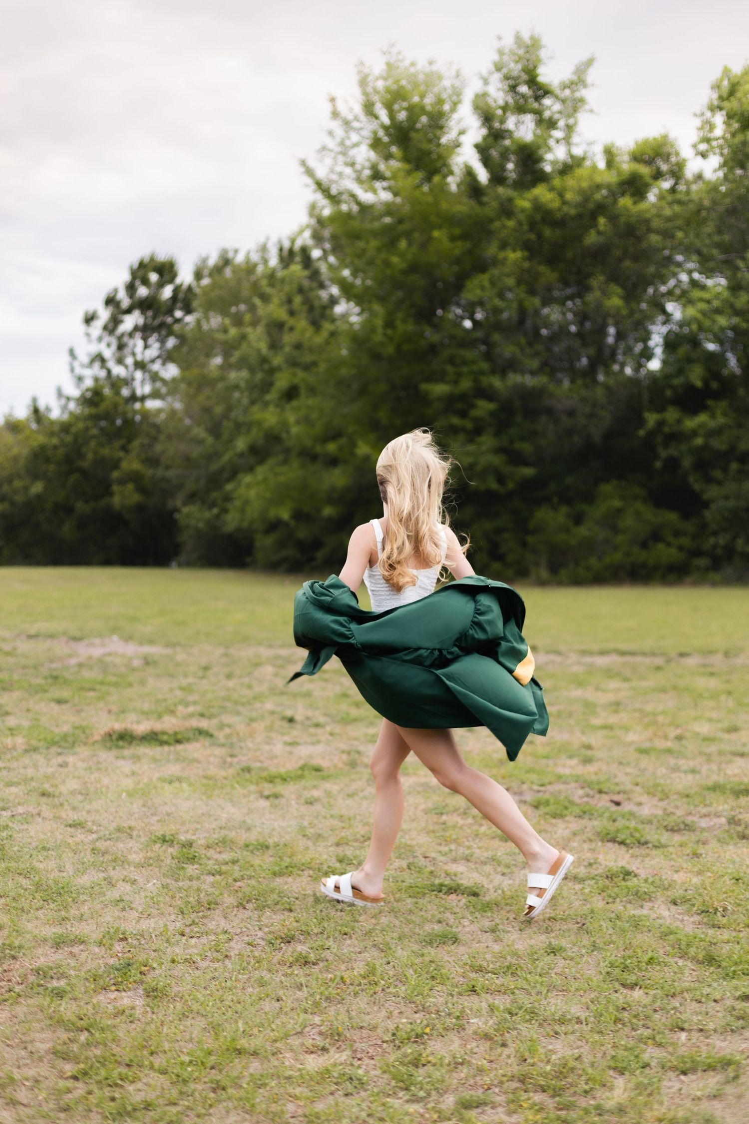 Blonde graduate twirling in green graduation gown over white dress in a grassy field with trees.