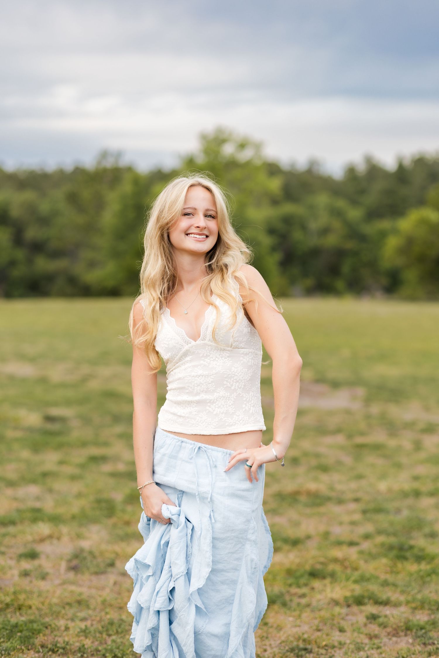 Smiling blonde woman in white crop top and light blue skirt posing in an open grassy field.