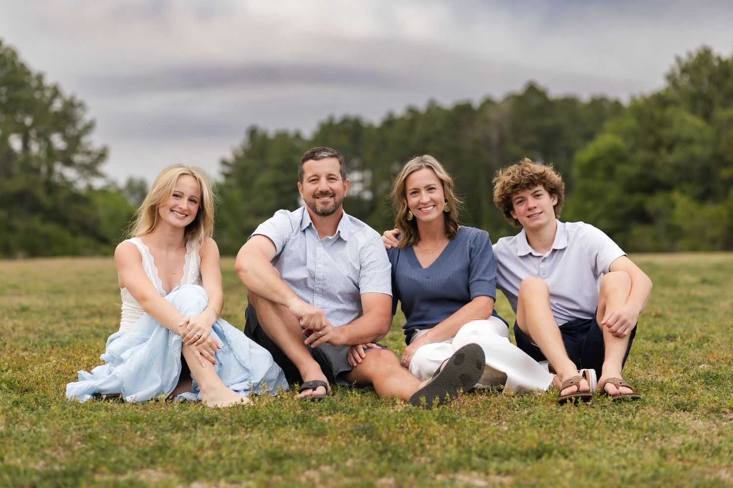 Happy family of four sitting on green grass outdoors, smiling together in a casual portrait setting.