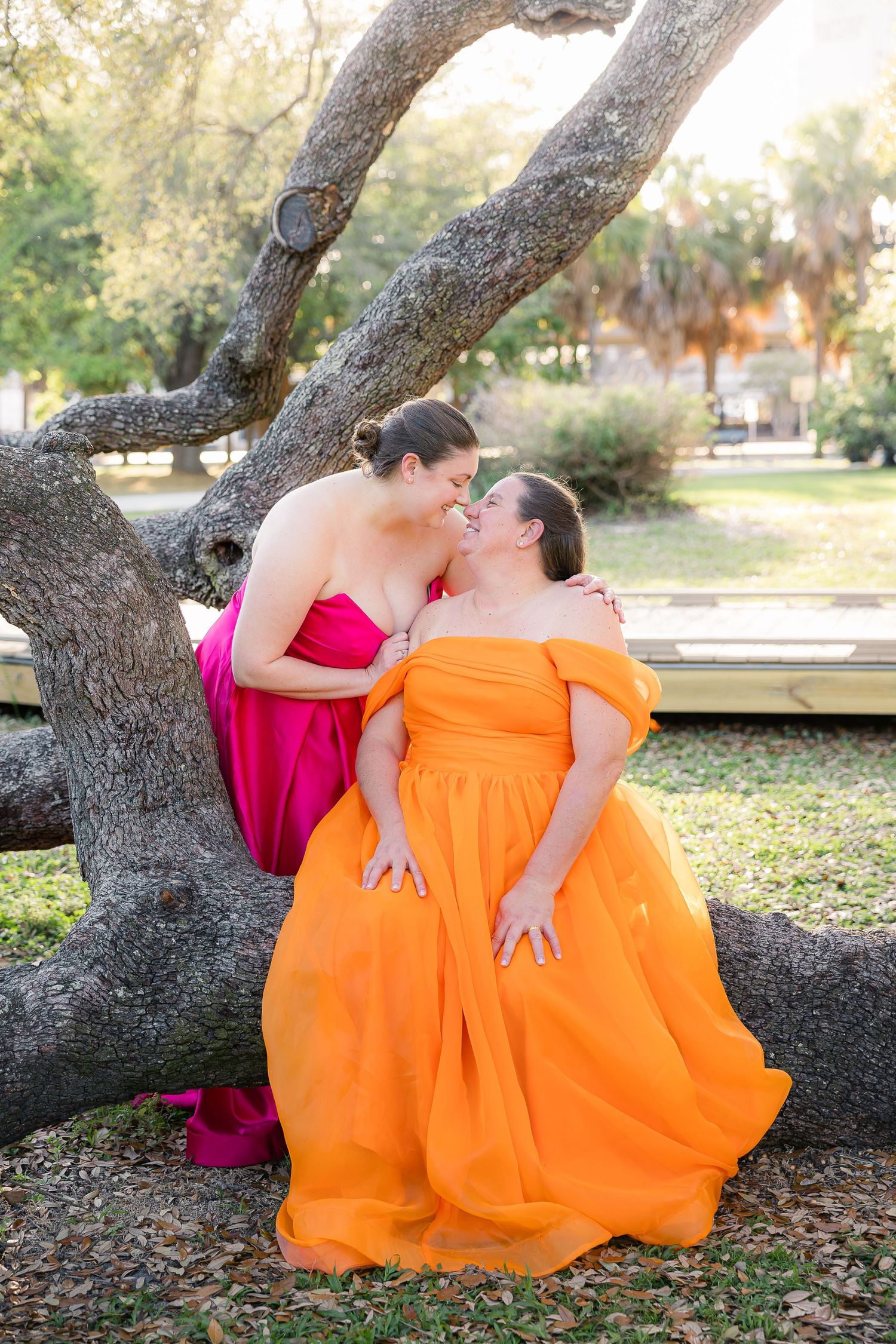 Two brides in vibrant pink and orange gowns share a romantic moment leaning against Treaty Oak in Jacksonville, Florida after ceremony.