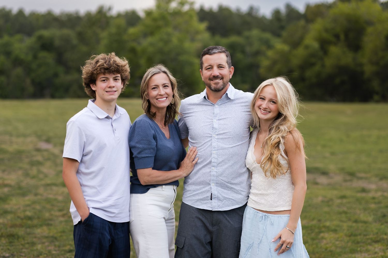 Family of four smiling outdoors in a green field, featuring two teenagers and their parents in casual attire.