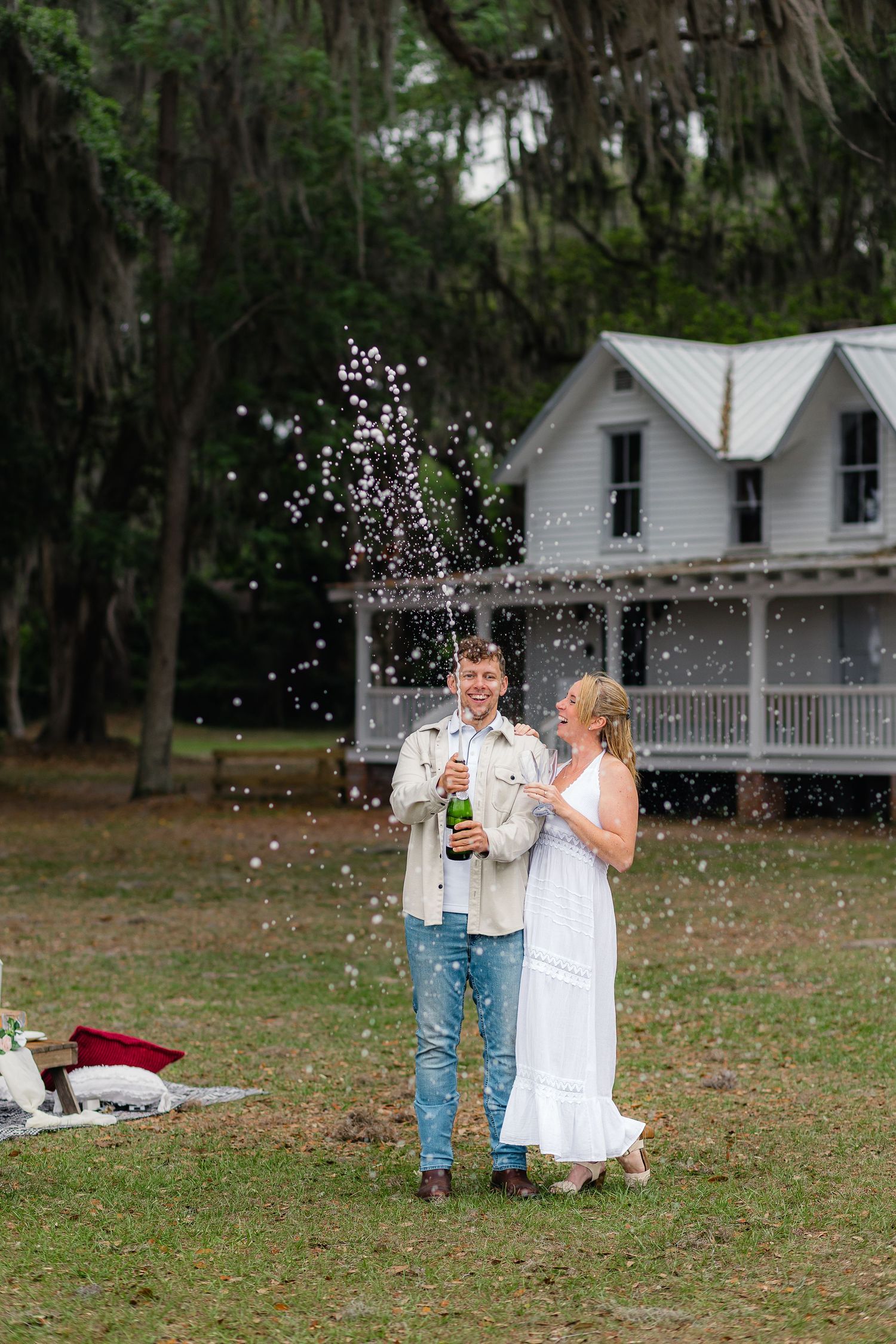 Couple celebrating engagement by popping champagne bottle at Alpine Grove Park under Spanish moss-covered trees.