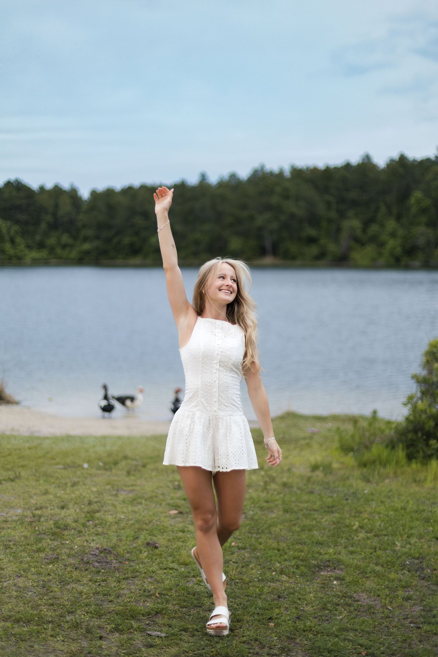 Happy blonde woman in white dress raising her arm near a peaceful lakeside setting.