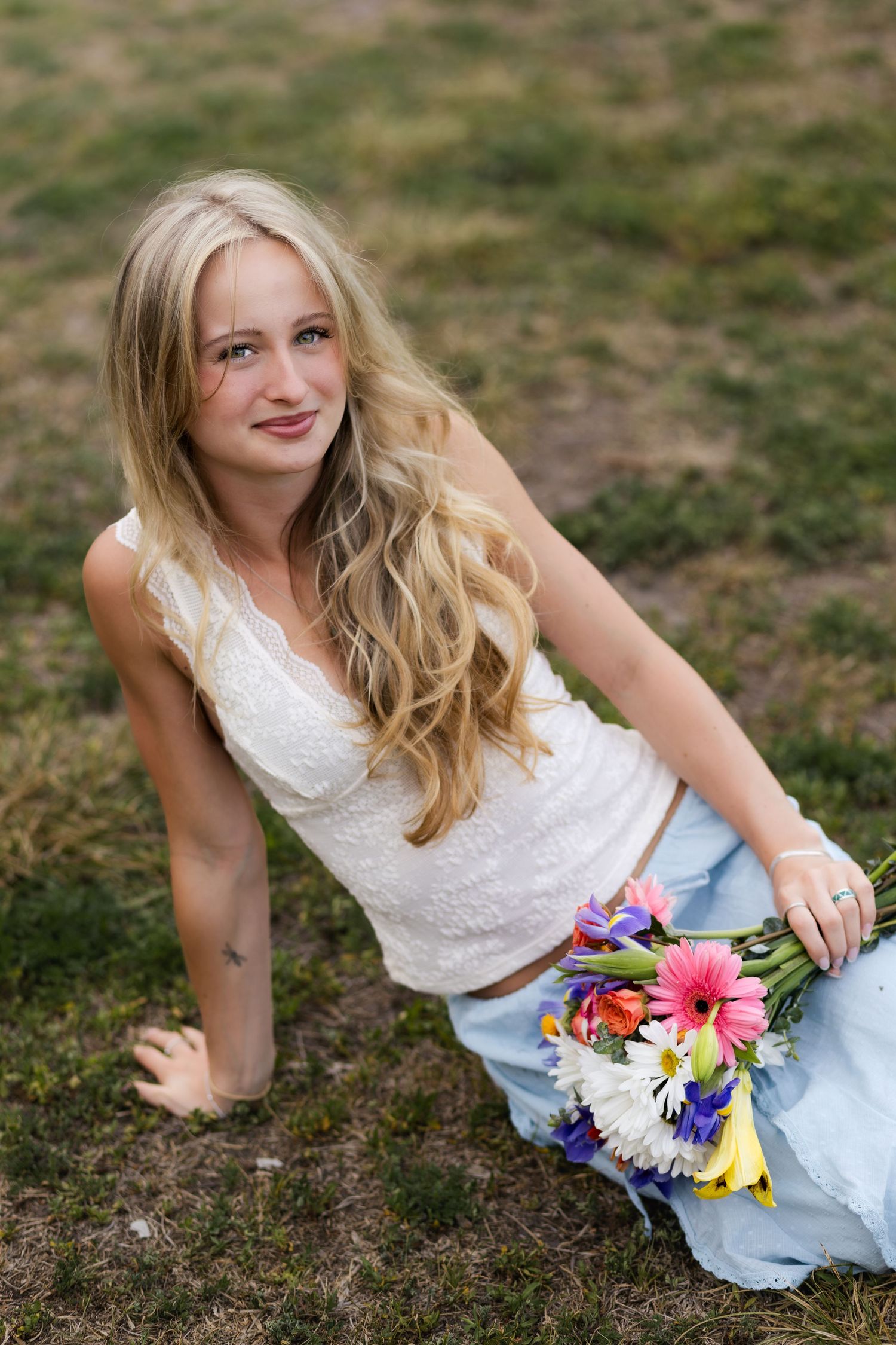 Smiling blonde woman sitting on grass holding colorful flower bouquet wearing white top and blue skirt.