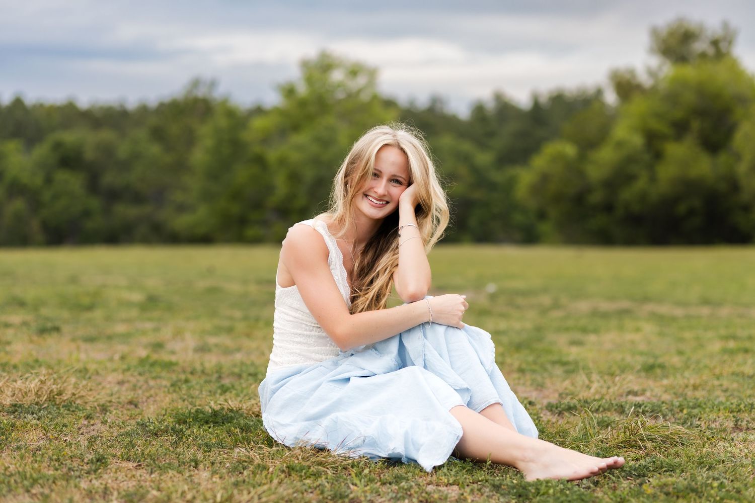 Smiling blonde woman in white top and light blue skirt sitting barefoot on green grass in an open field.