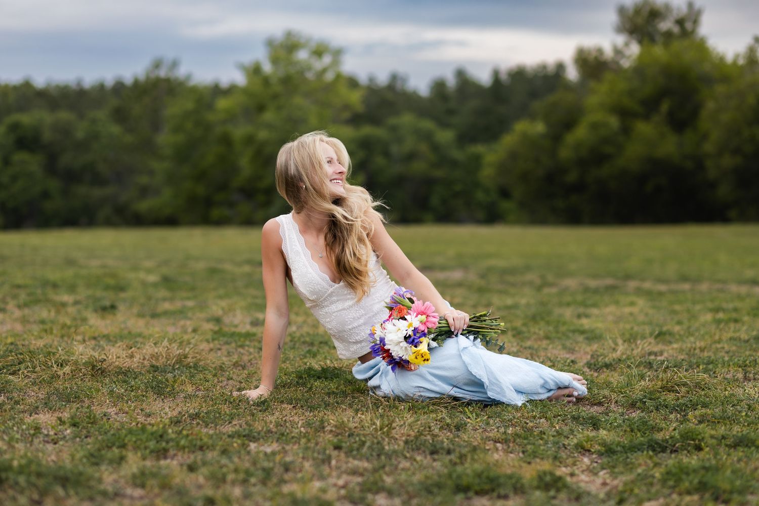 Blonde bride in white dress sitting on green grass field holding colorful bouquet of flowers outdoors.