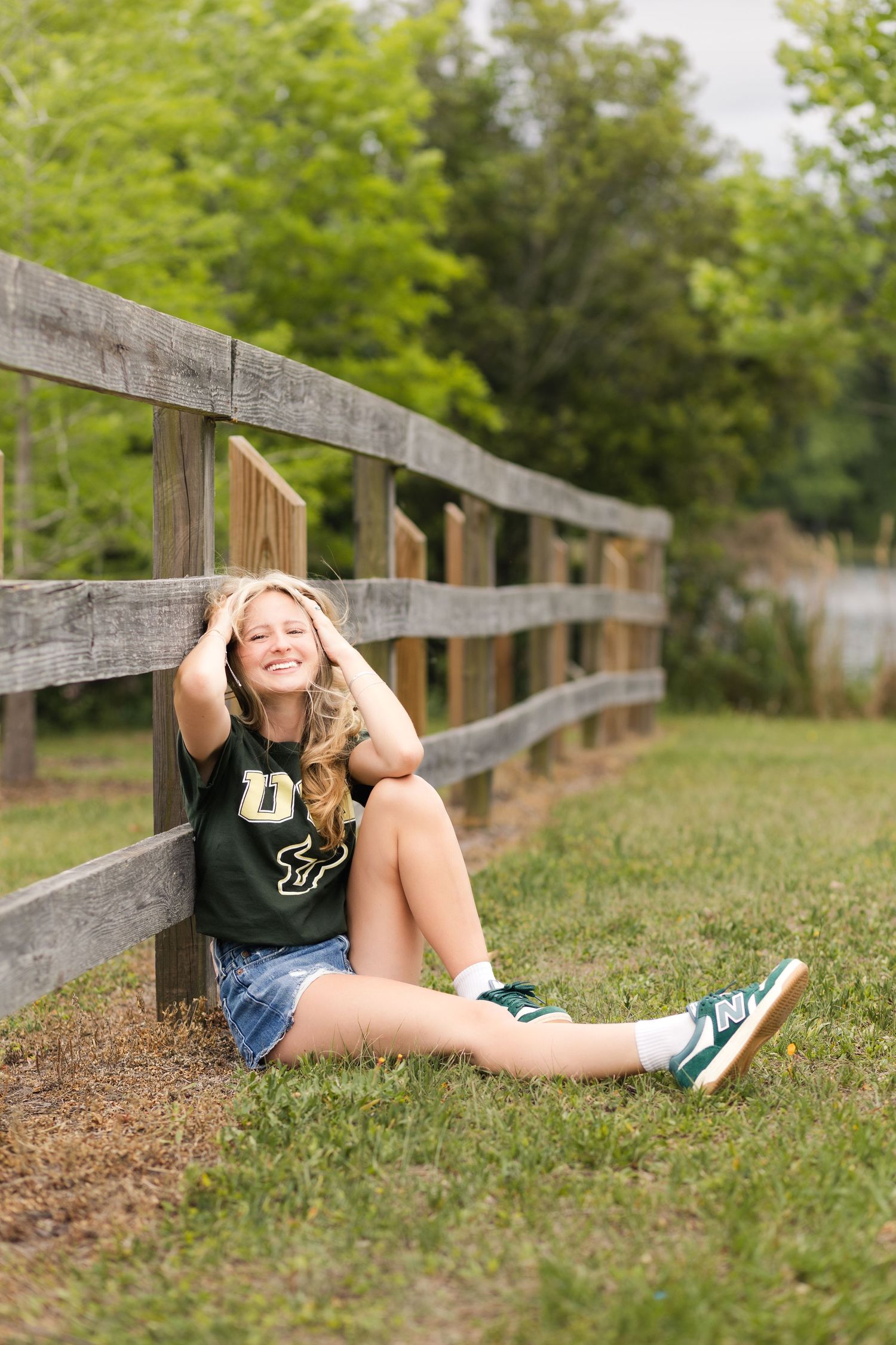 Laughing blonde teen in USF jersey and denim shorts sits against wooden fence in green park.