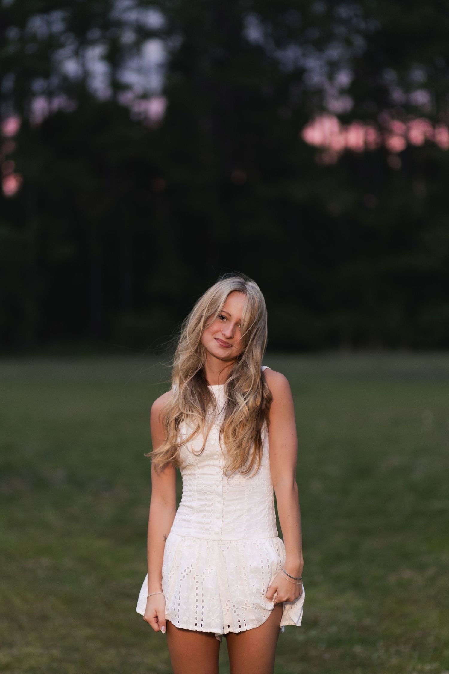 Young blonde woman in white mini dress stands in field at dusk with pink sunset sky behind her.