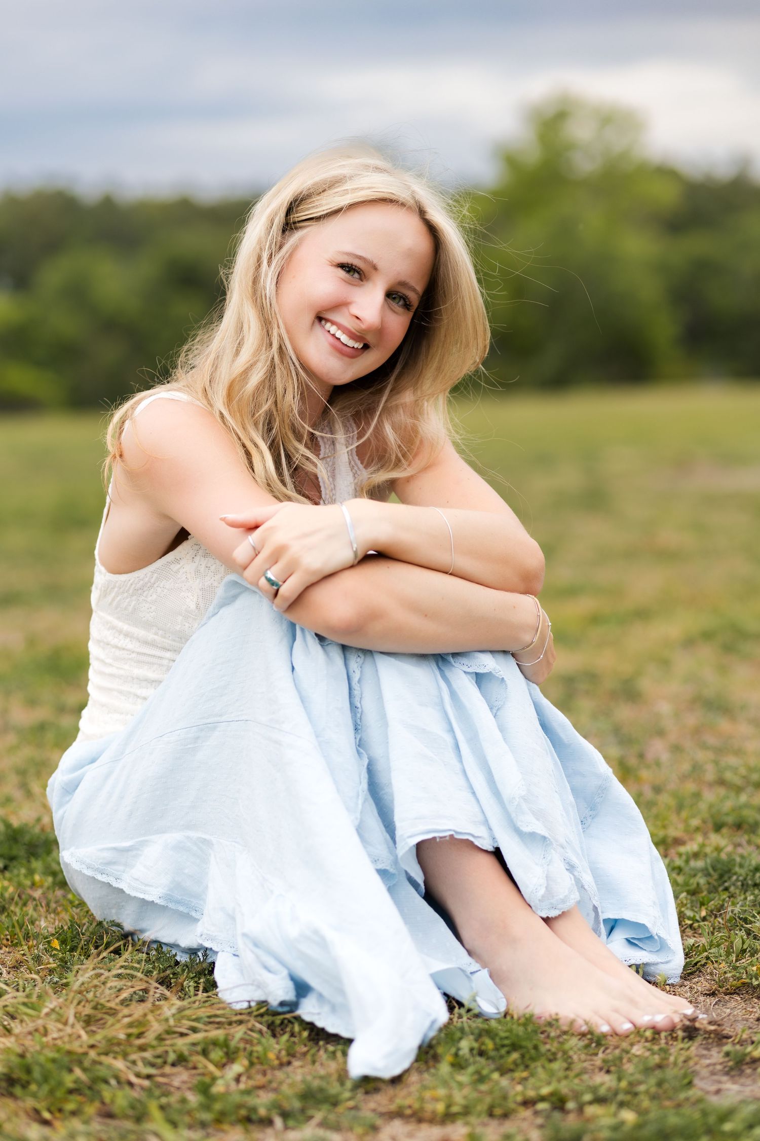 Smiling blonde woman in white top and light blue skirt sitting barefoot on grass in an outdoor field.