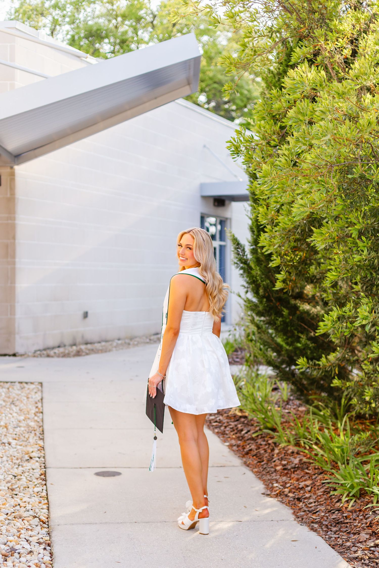 Girl woman walks along sidewalk holding graduation cap from Jacksonville Univeristy for senior portraits.