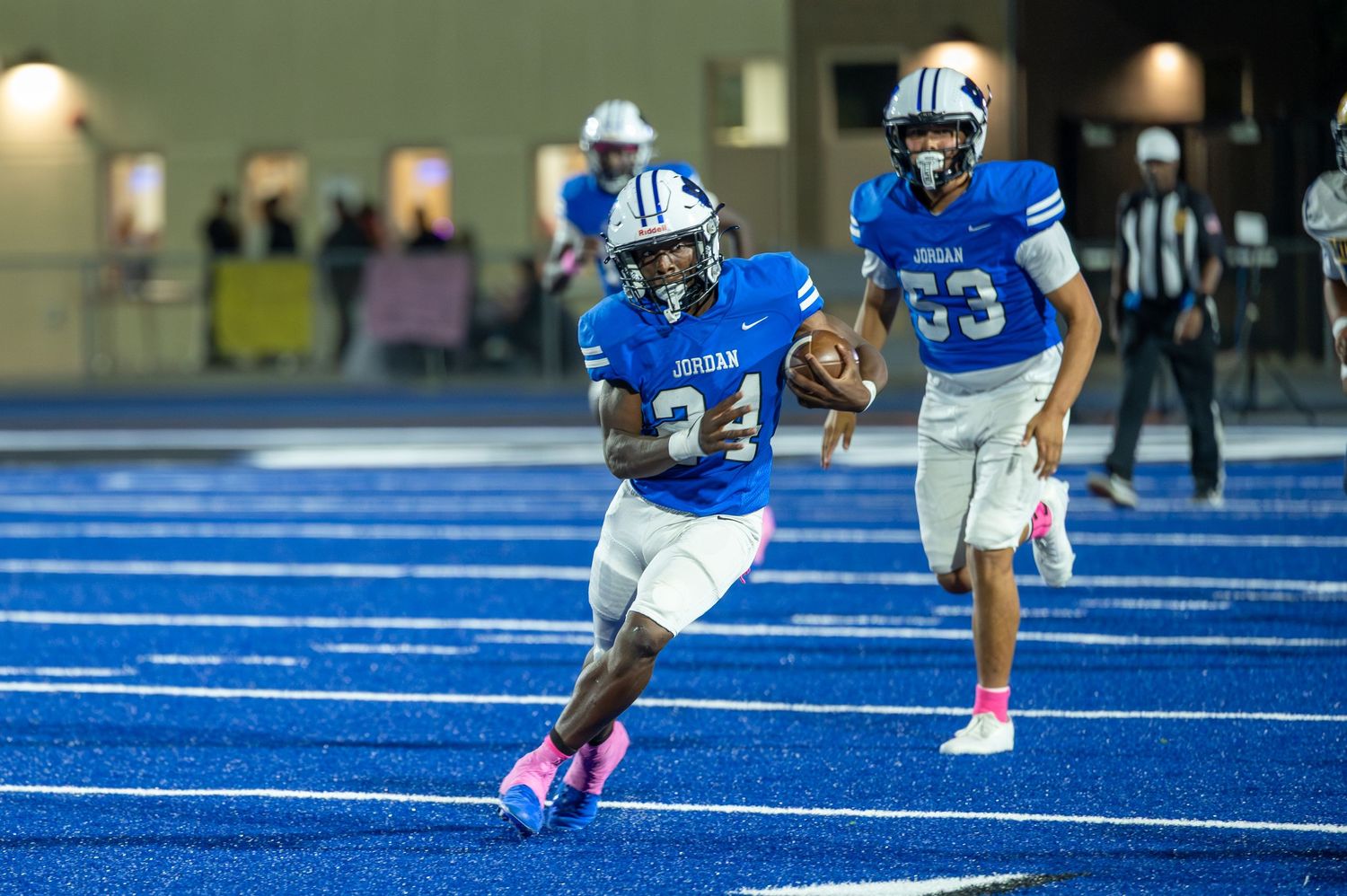 Football player #24 in blue Jordan jersey runs with ball on blue turf during night game.