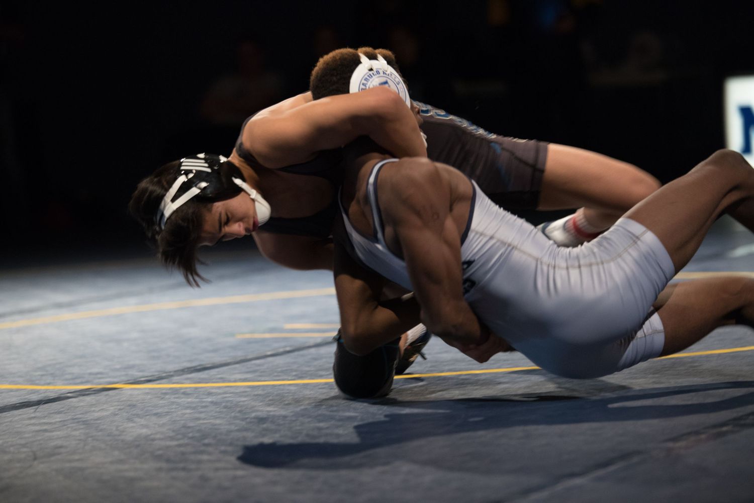 Two wrestlers grapple intensely on mat during competitive wrestling match under bright lights.