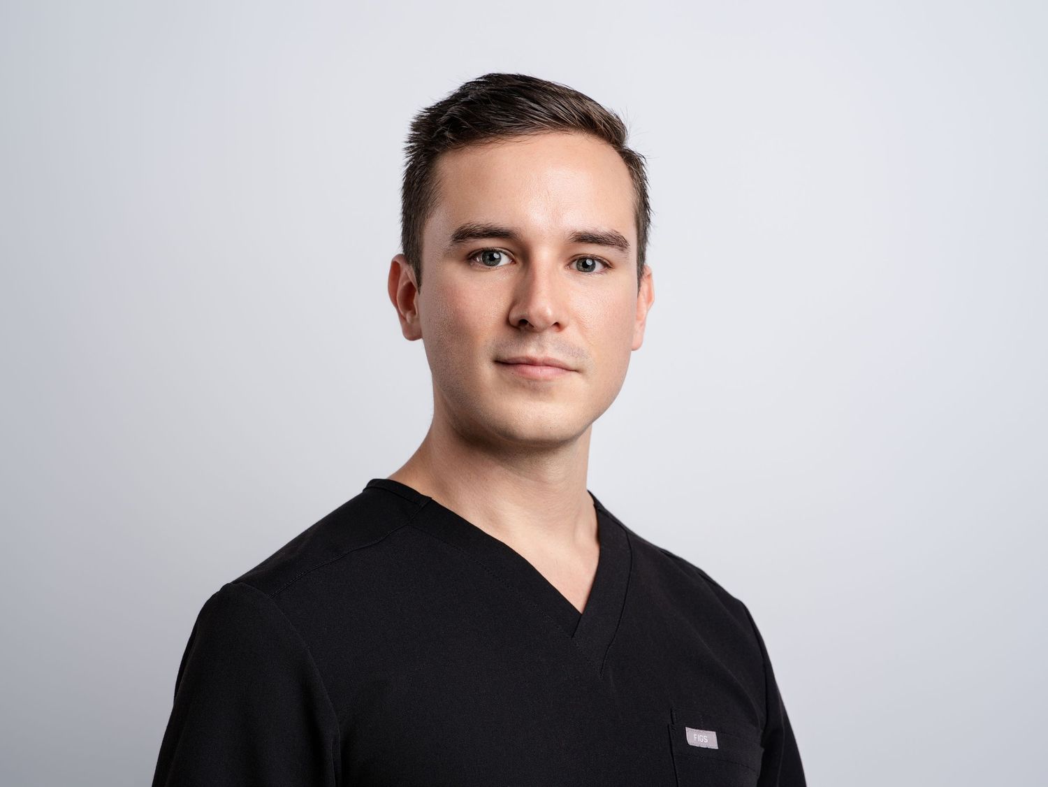 Young male healthcare professional in black scrubs poses for a clean studio headshot against a gray background.