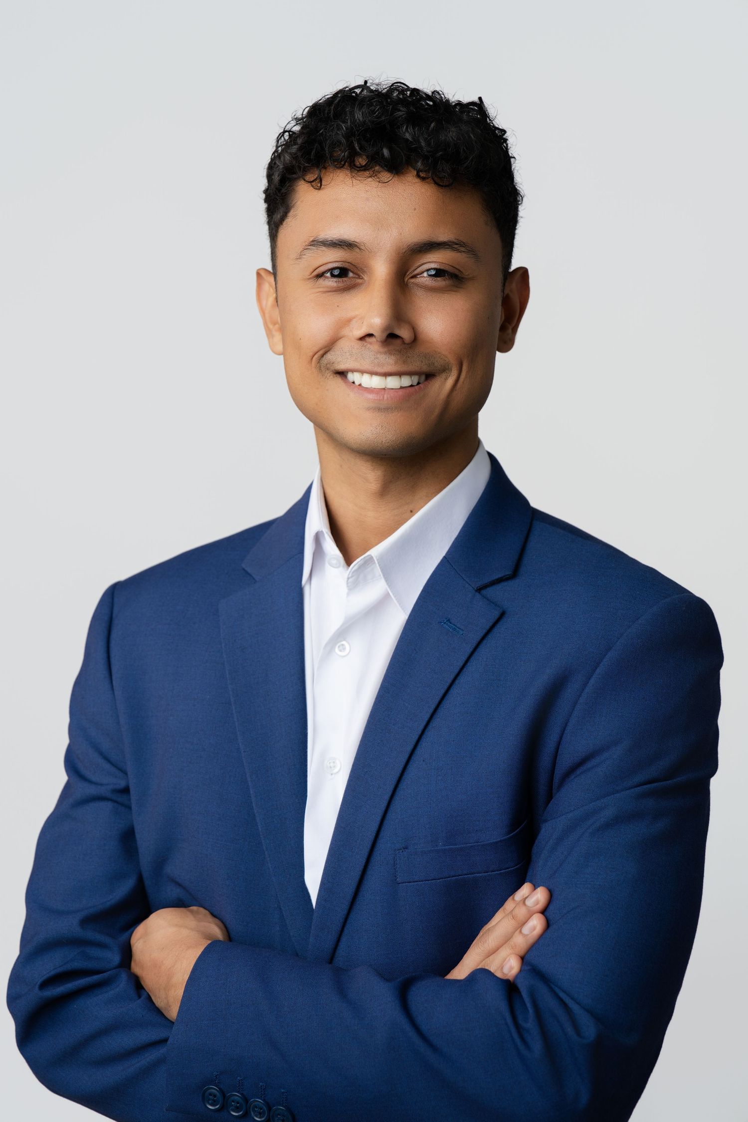 Confident young man in a navy suit smiles with arms crossed in a professional studio portrait.