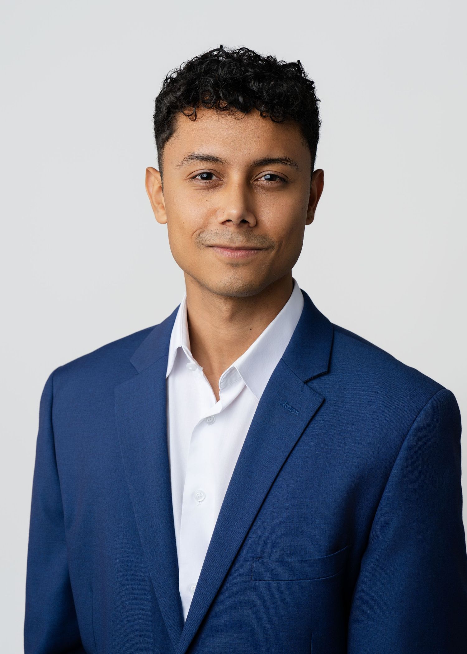 Young man in a navy blazer poses for a clean, professional headshot against a light gray background.