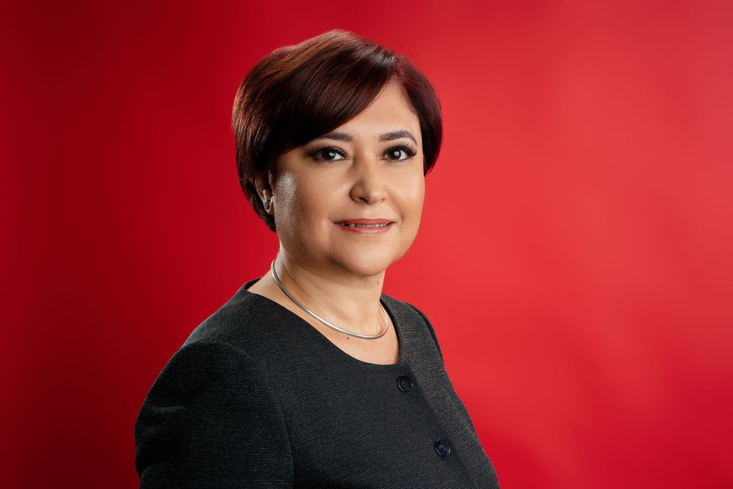 Professional woman with short auburn hair wearing a gray blazer poses against a bold red background.