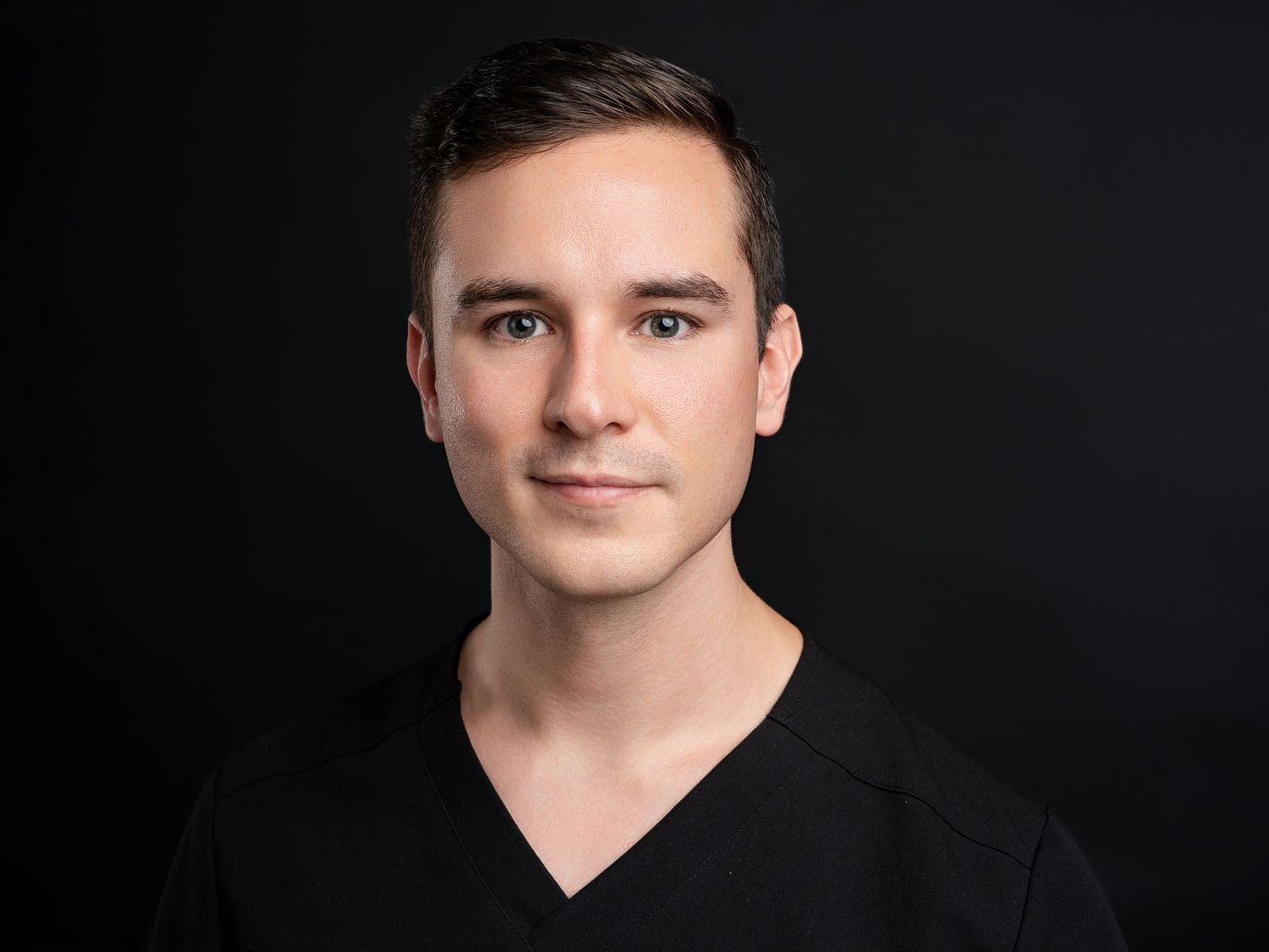 Professional headshot of a young man in a black v-neck top against a dark background.