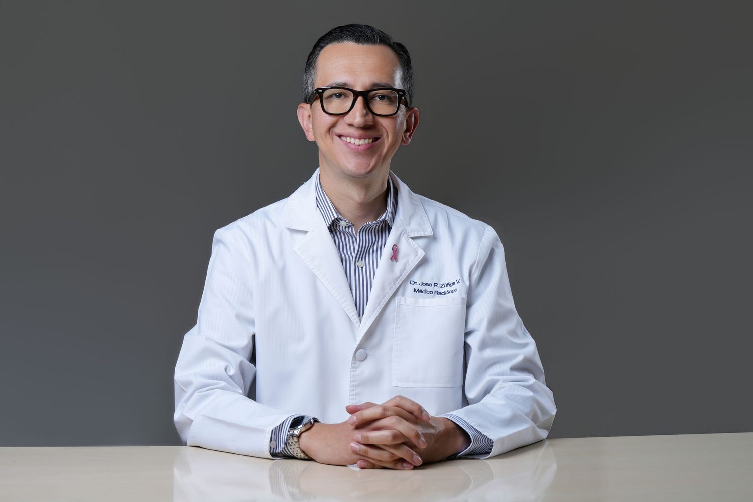 Professional portrait of a smiling man in a white lab coat seated at a table against a gray background.