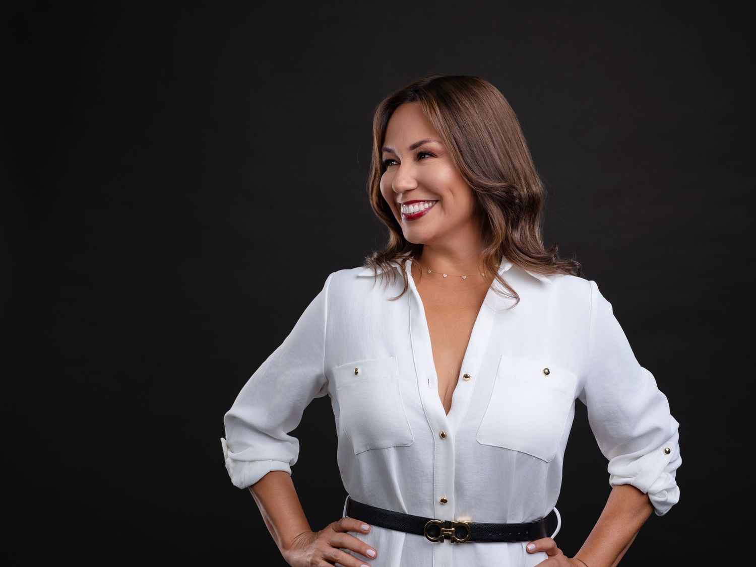 Confident woman in a white button-up blouse with a black belt poses against a dark background.