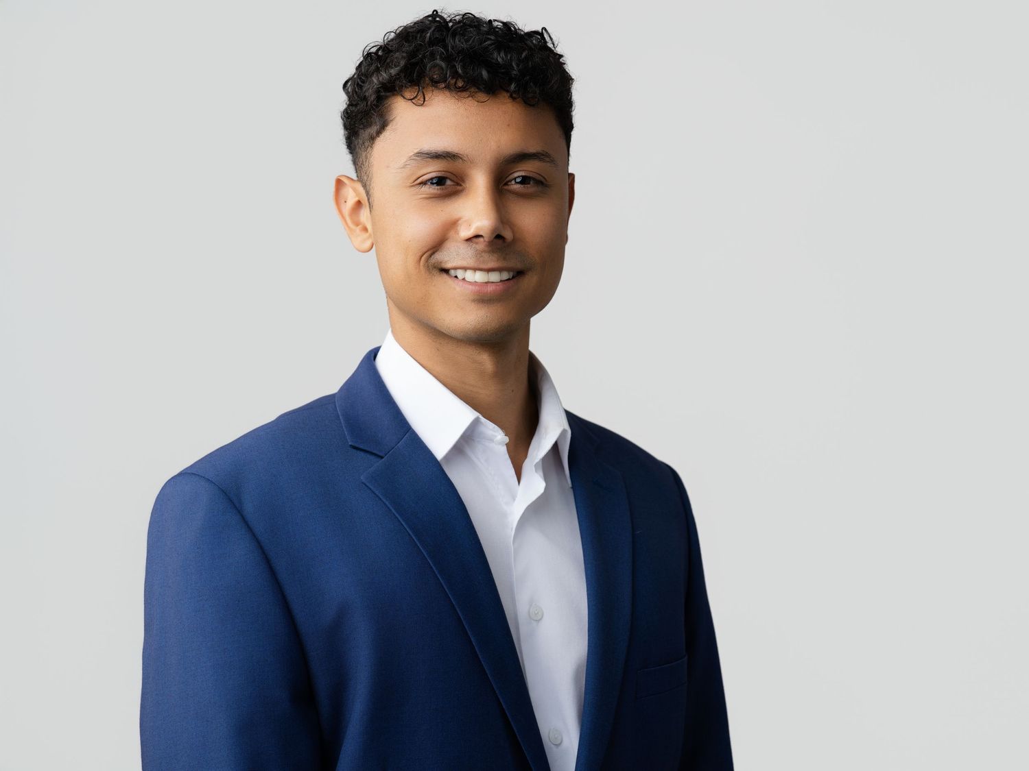 Smiling young man in a navy blue blazer poses for a professional headshot on a gray background.