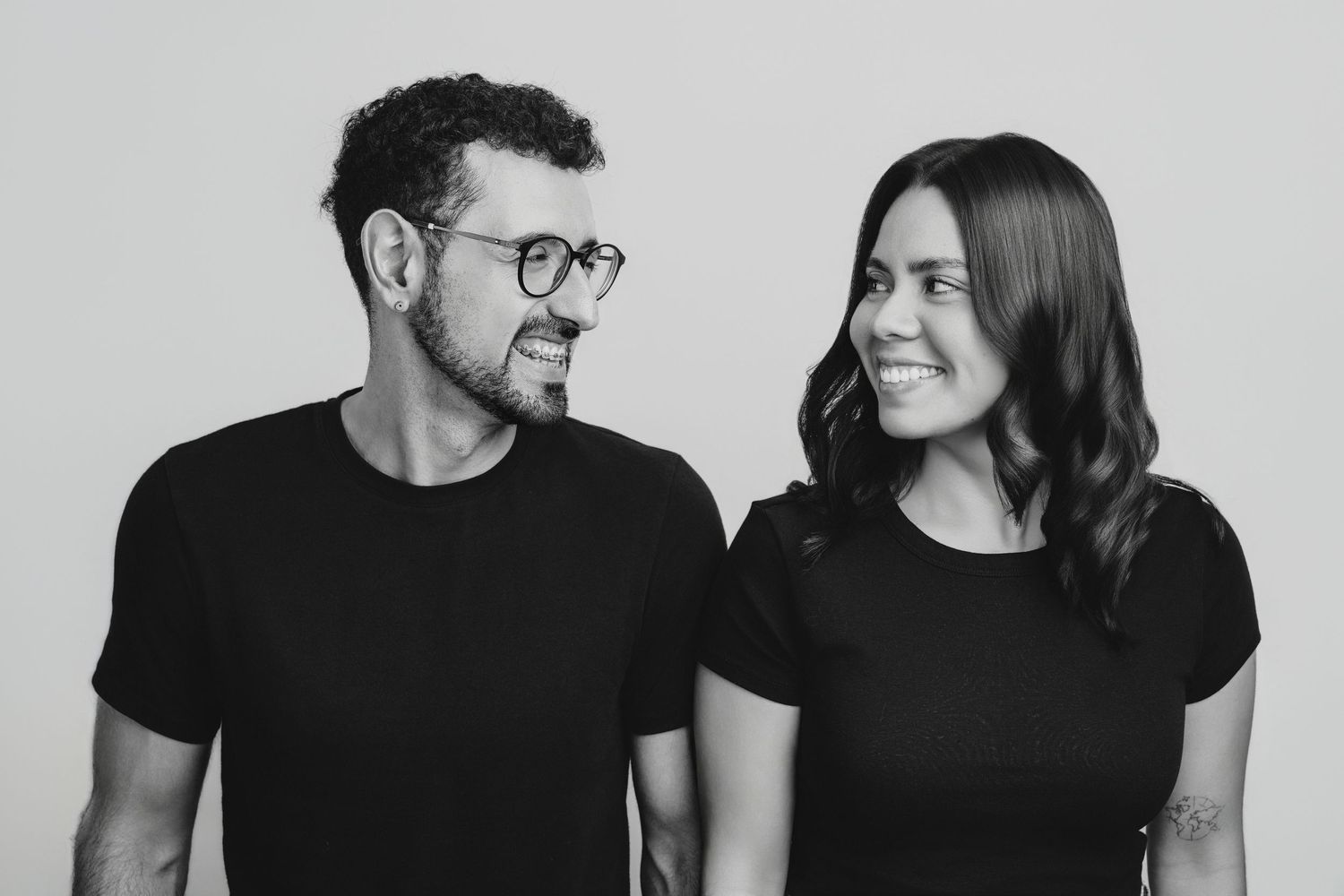 Black and white photo of a smiling man with glasses and a woman in black t-shirts looking at each other.