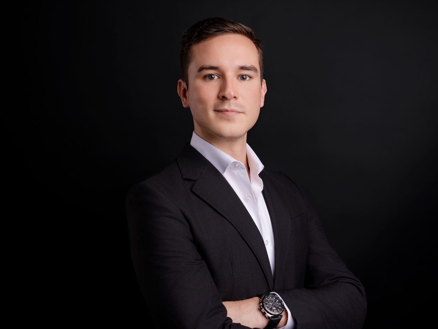 Professional headshot of a young man in a dark suit with arms crossed against a black background.