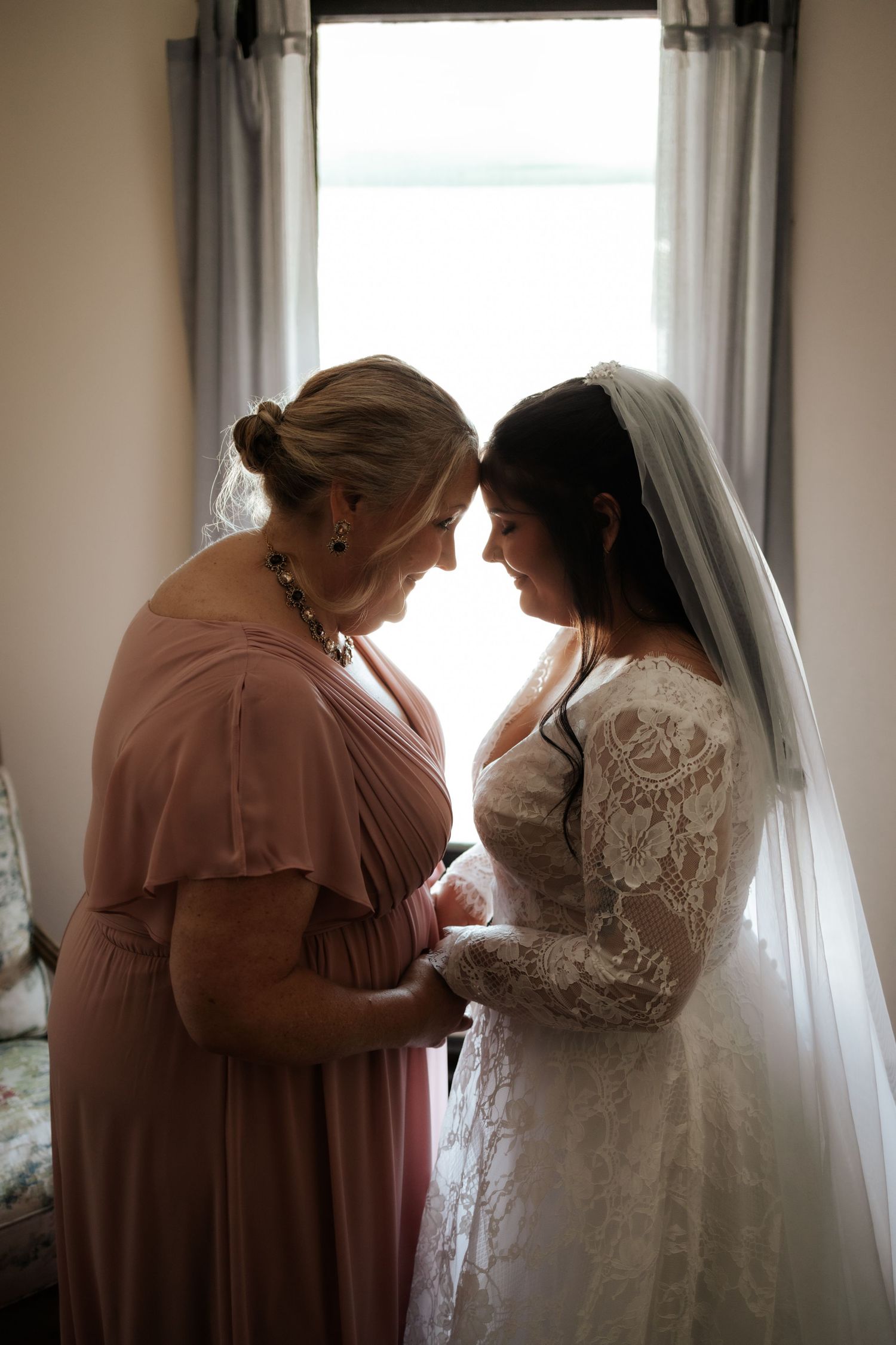 Bride in lace gown and veil shares tender forehead-to-forehead moment with bridesmaid in pink dress by window.