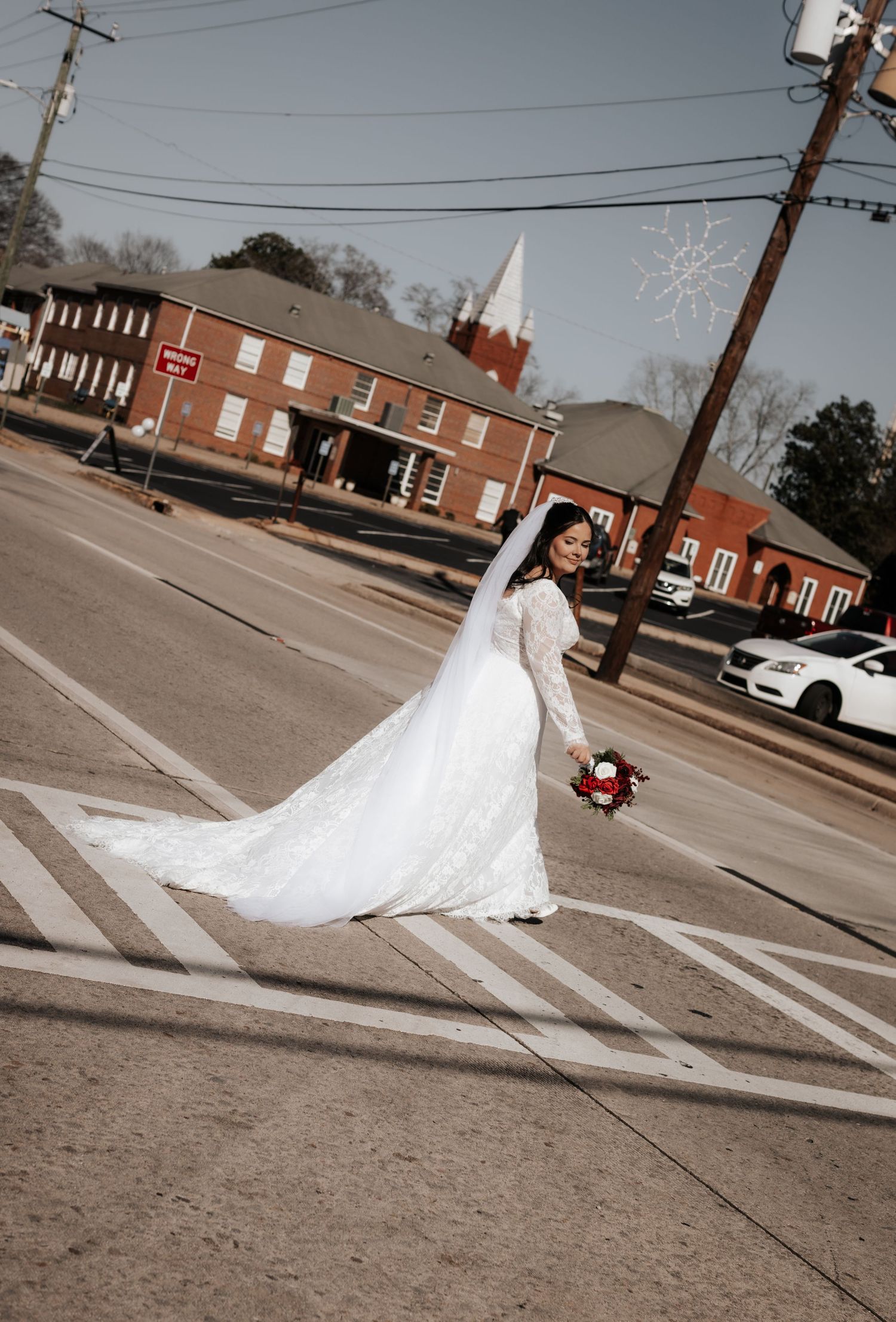 Bride in elegant white lace gown with long train and veil crosses a street intersection holding a red bouquet.