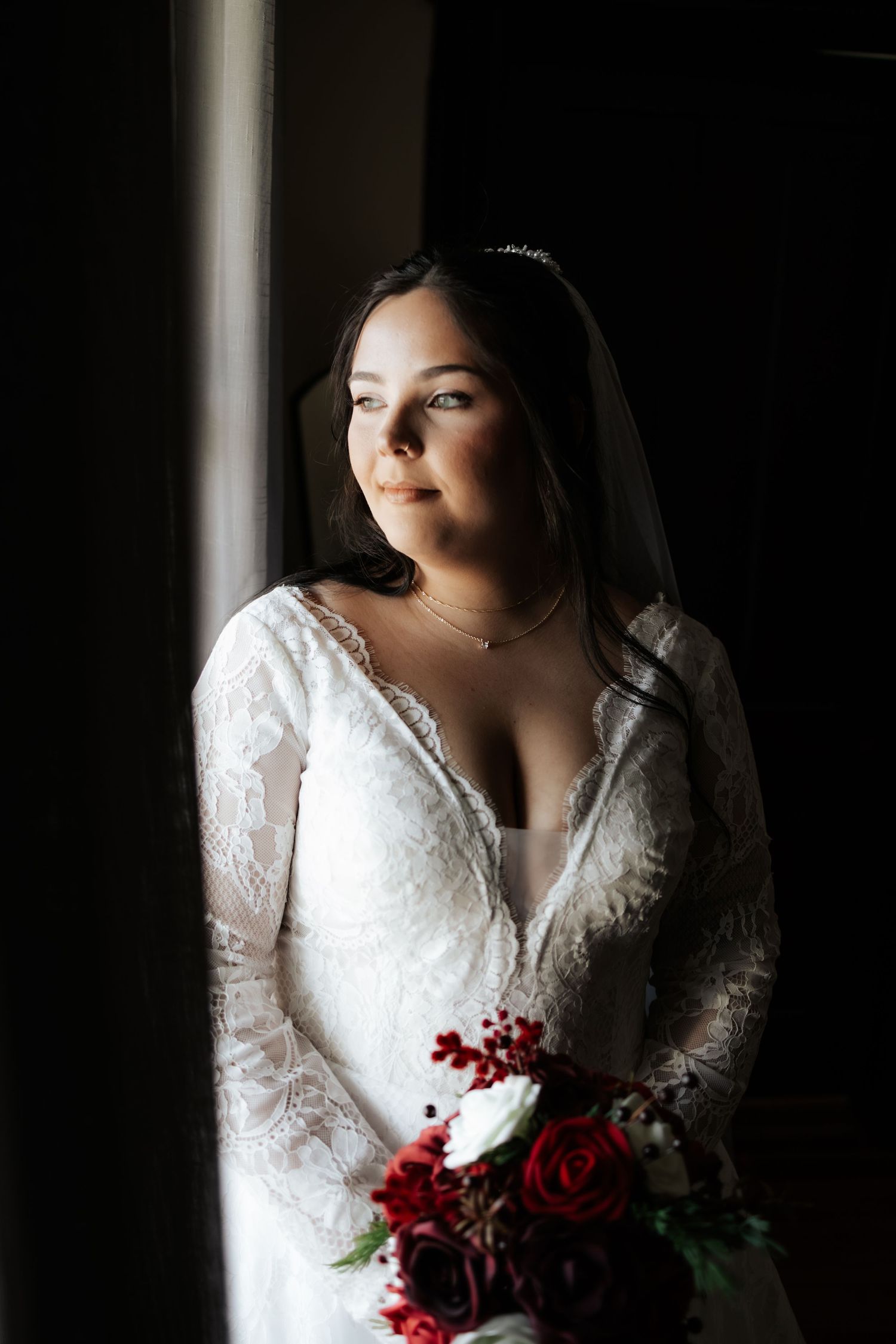 Bride in lace long-sleeve wedding dress holding red rose bouquet, standing by window in dramatic lighting.