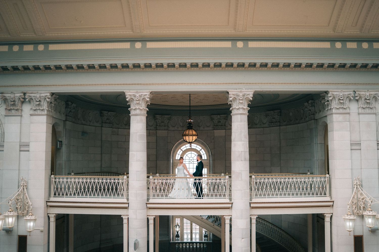Bride standing alone on a grand balcony inside an elegant historic venue with tall columns and ornate architecture,documentary wedding photo