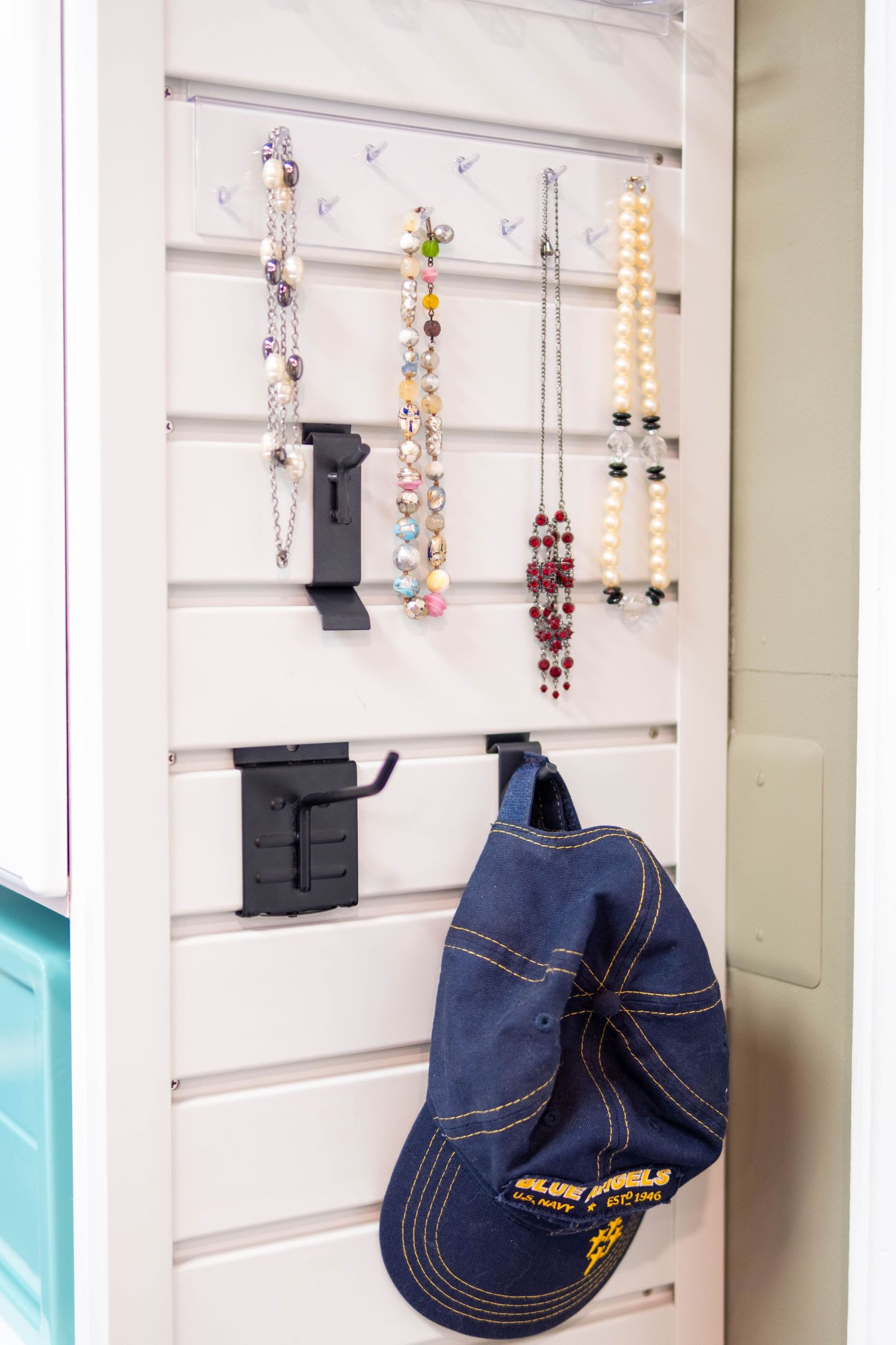 White pegboard organizer displaying colorful beaded necklaces, pearl strands, and a navy Blue Angels baseball cap on hooks.