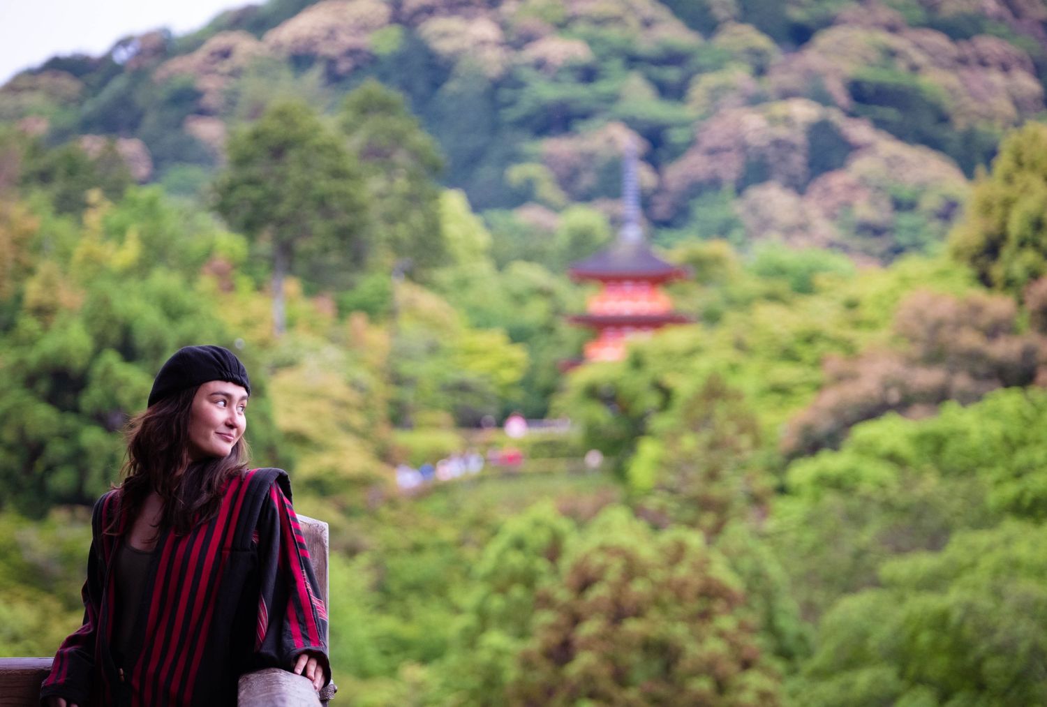 A smiling woman in a striped jacket sits outdoors with a lush green hillside and pagoda behind her.